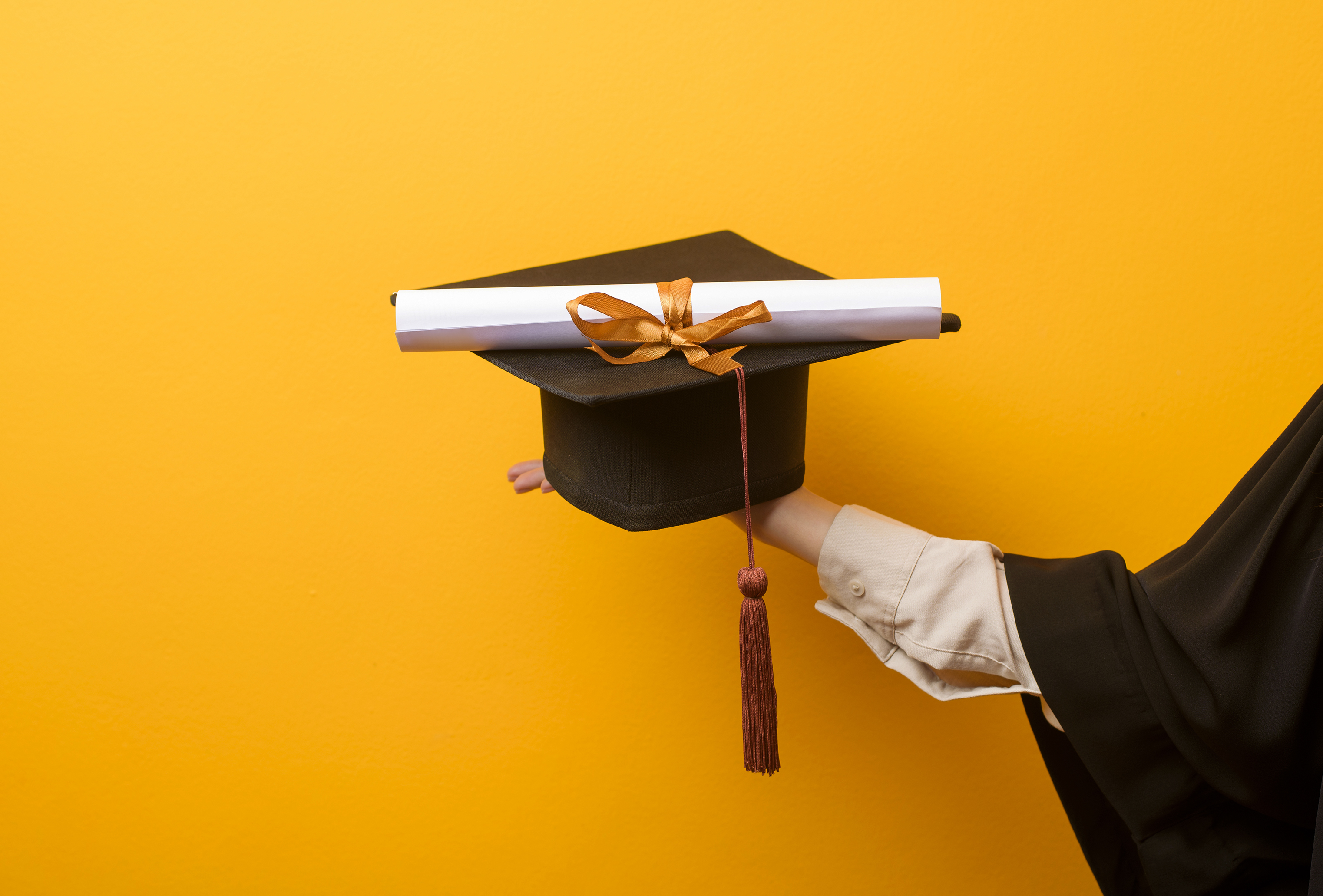 Close up of woman hand in graduation gown is holding graduation cap and certificate on yellow background 